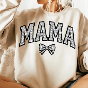 Könnte beinhalten: Beiges Sweatshirt mit dem Wort "MAMA" in großen, funkelnden, diamantartigen Buchstaben, schwarz umrandet. Eine Schleife mit ähnlichen funkelnden Details befindet sich darunter. Das Sweatshirt ist ein lässiger, bequemer Stil.