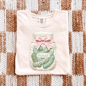 A white t-shirt with a pink bow and a design of pickles in a jar, laid out on a checkered background.