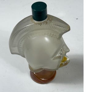 May include: A vintage perfume bottle shaped like a Roman soldier's head. The bottle is frosted glass with a dark green cap and a brown base. The bottle contains a yellow liquid.