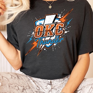 Puede incluir: Camiseta gris oscuro con el texto "OKC Thunder" en naranja y azul, con un diseño temático de baloncesto. El diseño incluye un rayo y una forma de escudo, con un estampado de leopardo en las letras.
