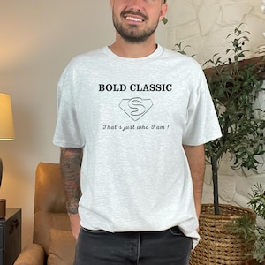 Può includere: T-shirt grigio chiaro con la scritta "BOLD CLASSIC" sopra una grafica a forma di diamante con la lettera "S" all'interno. Sotto, la frase "That's just who I am!"