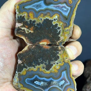 Puede incluir: Una losa de ágata pulida con bandas intrincadas en tonos azul, amarillo, naranja y marrón oscuro. La piedra muestra un patrón orgánico complejo con capas concéntricas y una formación central oscura de tipo dendrítico en la estructura mineral.
