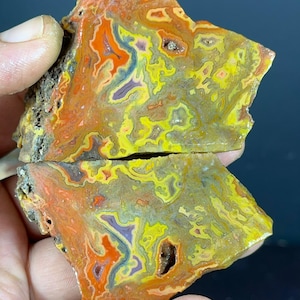 Puede incluir: Una losa de ágata pulida cortada en dos piezas, con bandas intrincadas en tonos vibrantes de naranja, amarillo brillante y morado apagado. La piedra muestra una forma natural e irregular con pequeñas cavidades huecas que contienen cristales.