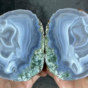 Op de afbeelding: Twee helften van een gepolijste agaatsteen, met concentrische banden van blauw en wit. De randen hebben een ruwe, groen en goudkleurige textuur. De steen wordt in twee handen gehouden, wat de natuurlijke schoonheid benadrukt.