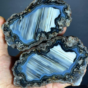 Può includere: Due fette di geode di agata lucidata con un nucleo centrale di bande verticali bianche e grigie, circondate da anelli concentrici di strati blu traslucidi e grigio antracite, con una crosta minerale esterna ruvida, strutturata e frastagliata.