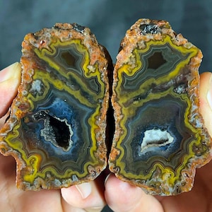 May include: A split agate geode featuring concentric bands of mustard yellow, deep blue, and rust orange. The interior displays a central crystalline cavity with dark and white mineral formations, surrounded by intricate, wavy, multi-coloured layers.