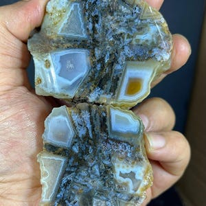 Op de afbeelding: Een gepolijste agaatplak met een geometrisch patroon van doorschijnende witte en lichtblauwe kristallijne secties, gescheiden door donkerbruine, grillige minerale aders. De steen heeft een onregelmatige, ruwe buitenrand.