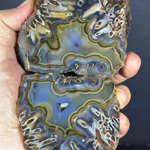 Può includere: Una lastra di agata lucidata caratterizzata da complesse bande concentriche nei toni del blu, giallo e marrone. La pietra presenta aree traslucide con inclusioni cristalline e bordi esterni frastagliati di colore marrone scuro.