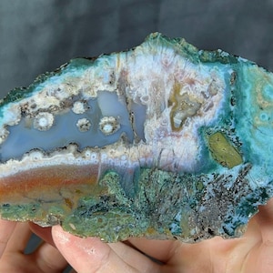 May include: A polished agate stone cross-section, displaying a range of colours and patterns. The stone has bands of blue, green, white, orange, and brown, with a crystalline structure. The shape is irregular, with a natural, organic appearance.
