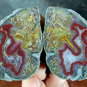 May include: Two halves of a polished agate stone, displaying a vibrant array of natural colours. The stone features a mix of red, yellow, and blue hues, with intricate patterns and textures. The agate is held in a person's hand.
