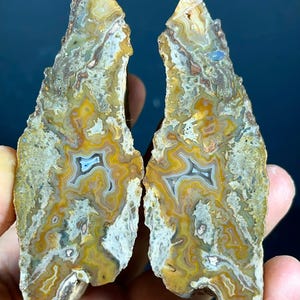 Puede incluir: Dos mitades a juego de una rodaja de geoda de ágata pulida con intrincadas bandas concéntricas en tonos amarillo mostaza, crema, gris y marrón. La piedra muestra un borde exterior natural dentado y una superficie interior lisa y plana.
