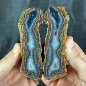 Könnte beinhalten: Zwei Hälften eines polierten Achats, die konzentrische Farbbänder zeigen. Der Stein weist Schichten aus blauen, weißen, braunen und beigen Farbtönen auf. Die rauen Kanten kontrastieren mit dem glatten, polierten Inneren und zeigen komplizierte Muster.