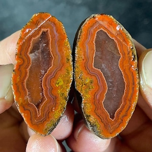 May include: Two halves of a polished agate stone, displaying concentric bands of orange, red, and brown. Each piece has a crystalline cavity at its centre. The stone's surface is glossy, accentuating the natural patterns and colours.