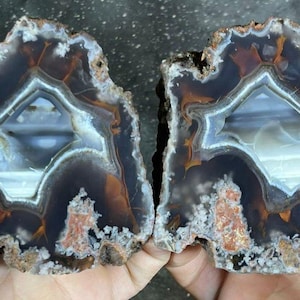 May include: Two halves of a polished agate stone, displaying a natural, layered pattern. The stone has a dark brown outer edge, leading to lighter brown and white bands, with a central, crystalline cavity. The rough edges are visible.