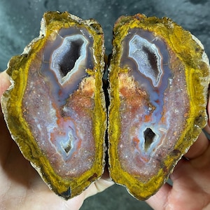 Può includere: Due metà di una pietra d'agata lucidata, che mostra una formazione di geode naturale. La pietra presenta una vibrante tavolozza di colori giallo, arancione, viola e bianco, con un centro cristallino. L'esterno ruvido contrasta con l'interno liscio e lucido.