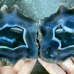 May include: Two halves of a polished agate stone, showcasing a striking blue and black pattern with white accents. The natural stone has a unique, organic shape, and is held in two hands.
