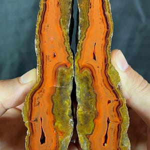 Puede incluir: Dos mitades de una piedra de ágata pulida, que muestran bandas vibrantes de color naranja, marrón y amarillo. La sección transversal de la piedra revela patrones intrincados y huecos naturales, sostenidos por manos sobre un fondo oscuro.