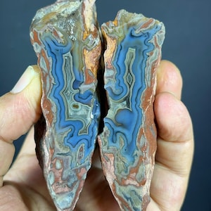 Op de afbeelding: Twee helften van een gespleten agaatgeode met ingewikkelde, golvende banden in tinten van diepblauw, roestoranje en crème. De steen vertoont een gepolijst, kristallijn interieur met een ruwe, getextureerde buitenrand en natuurlijke formaties.