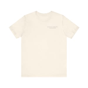 以下が含まれることがあります： クリーム色のTシャツ。黒で「QUIETLY STRONG」の文字が細い黒い線の上にプリントされています。半袖でクルーネック。生地は柔らかく、軽量に見えます。