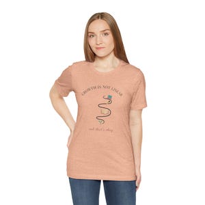 May include: Peach-colored t-shirt with the text "Growth is not linear" and "and that's okay" in a curved design. The graphic includes a book, a graduation cap, and a winding line. The shirt is made of a soft material.