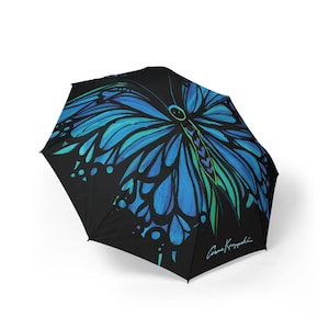 May include: Black umbrella featuring a large, vibrant blue butterfly design. The butterfly's wings have intricate patterns and gradients of blue and teal. The umbrella has a curved handle and a signature in the lower right corner.