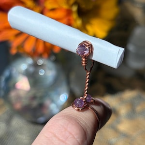May include: A copper wire ring featuring a twisted band design with two small, round, light purple amethyst beads at the base and a single matching amethyst bead at the top, which acts as a holder for a small, tightly rolled white paper scroll.