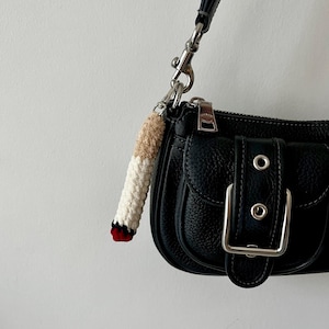 May include: A black textured leather handbag with a silver buckle and a crochet keychain shaped like a cigarette. The keychain features a tan filter, a white body, and a red and black tip, attached to the bag's silver metal hardware.