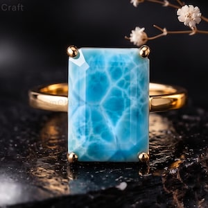 May include: A gold ring featuring a large, rectangular, light blue Larimar gemstone with a distinct white marbled pattern. The stone is held in a four-prong setting on a smooth, polished gold band, showcasing a vibrant sky-blue color.