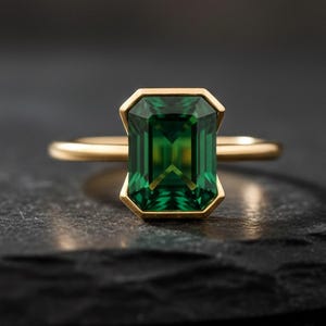 May include: A gold ring featuring a rectangular emerald-cut green gemstone set in a smooth bezel setting. The band is thin and polished, showcasing a clean design that highlights the deep green color and faceted clarity of the central stone.