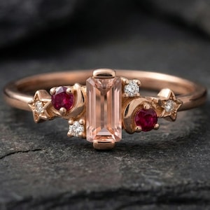 May include: Rose gold ring with a rectangular peach gemstone, ruby and star and moon accents. Small diamond accents are also present. The ring is set against a dark, textured background.