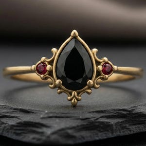 May include: A gold ring with a central, pear-cut black gemstone and two smaller red stones. The ring has an elaborate, vintage design with intricate filigree patterns. The ring's design is reminiscent of antique jewellery.