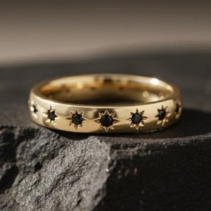 May include: A gold-colored ring with a series of black star-shaped accents. The ring is set on a dark, textured surface. The ring's design features a repeating pattern of stars around the band, creating a celestial theme.