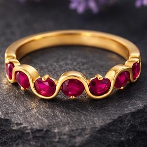 May include: A gold ring with oval-cut, vibrant red gemstones set in a repeating pattern. The ring is displayed on a dark gray, textured surface, with blurred purple flowers in the background. The ring's design is elegant and eye-catching.