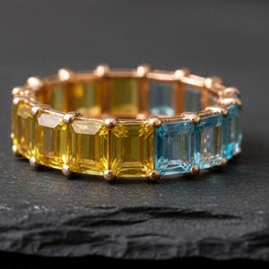 May include: A gold-toned ring featuring a band of alternating yellow and blue rectangular gemstones. The ring is set on a dark, textured surface, showcasing the vibrant colors of the stones. The gemstones are evenly spaced around the band.