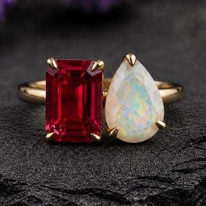 Könnte beinhalten: Ein Goldring mit zwei markanten Edelsteinen: einem rechteckigen, smaragdgeschliffenen tiefroten Stein und einem birnenförmigen, schillernden weißen Opal. Die Steine sind in einer Krappenfassung nebeneinander auf einer schmalen Goldschiene gefasst.