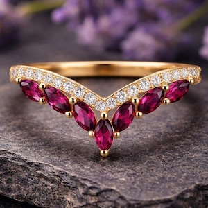 May include: A gold ring with a V-shaped design. The ring features a row of small, round, clear stones and a series of marquise-cut, deep red gemstones. The ring is set on a dark grey stone surface, with blurred purple flowers in the background.