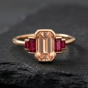 Puede incluir: Un anillo de oro con una gran gema rectangular de color melocotón en el centro, flanqueada por dos gemas rojas rectangulares más pequeñas a cada lado. El anillo está sobre una superficie oscura y texturizada.