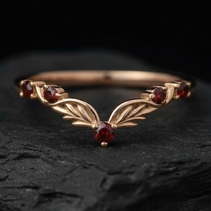 May include: A rose gold ring with a V-shaped design. The ring features small, round, red gemstones and leaf-like details. The ring is set against a dark background.