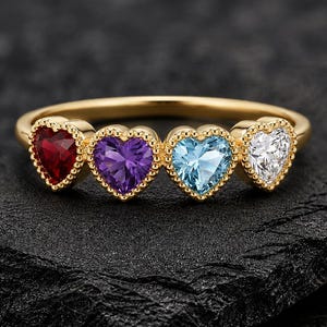Può includere: Un anello in oro con quattro pietre preziose a forma di cuore disposte in fila. Le pietre includono un rubino rosso intenso, un'ametista viola vibrante, un'acquamarina azzurra e un diamante bianco, incastonate in una montatura in oro.