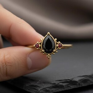 May include: A gold ring with a central, pear-cut black gemstone and two smaller red stones. The ring has an elaborate, vintage design, with a decorative setting.
