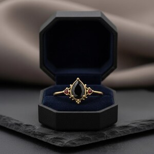 May include: A gold ring featuring a teardrop-shaped black gemstone and two small red stones. The ring is presented in a black, octagonal jewellery box with a blue velvet lining. The ring has an elaborate design.
