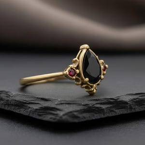 May include: A gold ring with a pear-cut black gemstone at its centre. Two small red gemstones flank the black stone. The ring has an ornate, vintage design, with a gold band and decorative detailing.