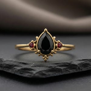 May include: A gold ring with a central pear-cut black gemstone and two smaller red gemstones. The ring has an elaborate, vintage design with intricate filigree. The ring is displayed on a dark, textured surface.