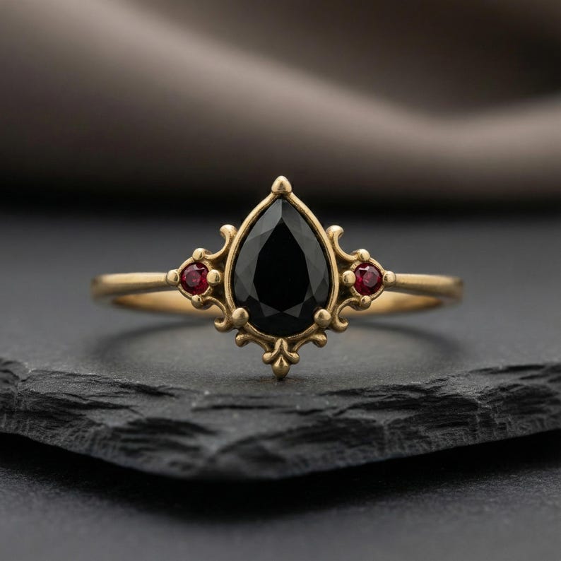 May include: A gold ring with a central pear-cut black gemstone and two smaller red gemstones. The ring has an elaborate, vintage design with intricate filigree. The ring is displayed on a dark, textured surface.