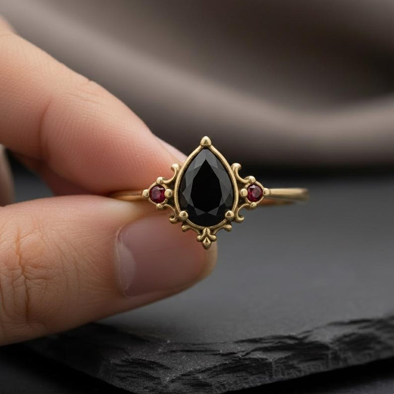 May include: A gold ring with a central, pear-cut black gemstone and two smaller red stones. The ring has an elaborate, vintage design, with a decorative setting.