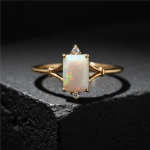 May include: A gold ring with a rectangular opal center stone and two small accent diamonds. The opal has a milky white base with flashes of color. The ring is set on a dark surface.