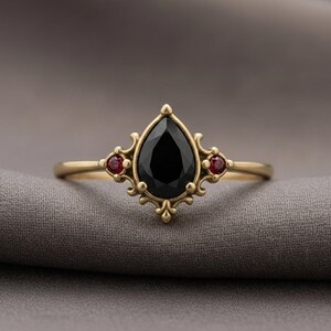 May include: A gold ring with a large, pear-cut black gemstone and two smaller red stones. The ring has an elaborate, vintage design. The ring's design is reminiscent of antique jewellery.