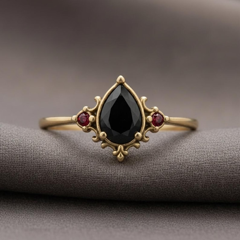 May include: A gold ring with a large, pear-cut black gemstone and two smaller red stones. The ring has an elaborate, vintage design. The ring's design is reminiscent of antique jewellery.