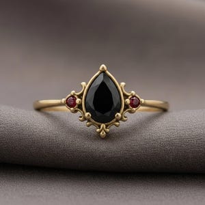 May include: A gold ring with a central, pear-cut black gemstone and two smaller red stones. The ring has an elaborate, vintage design. The ring's design is reminiscent of antique jewellery.
