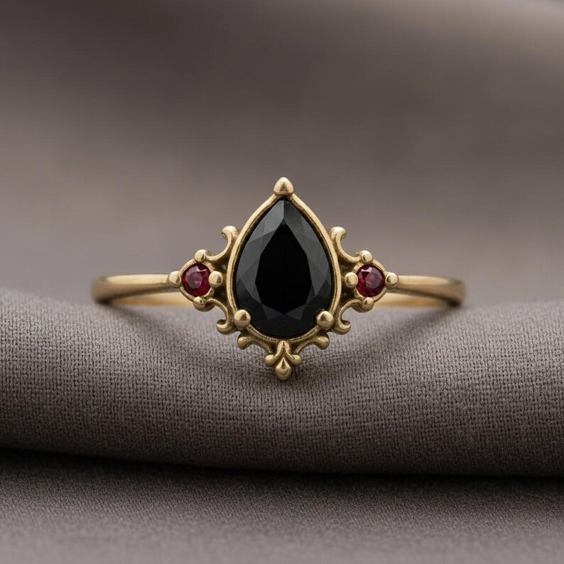 May include: A gold ring with a central, pear-cut black gemstone and two smaller red stones. The ring has an elaborate, vintage design. The ring's design is reminiscent of antique jewellery.